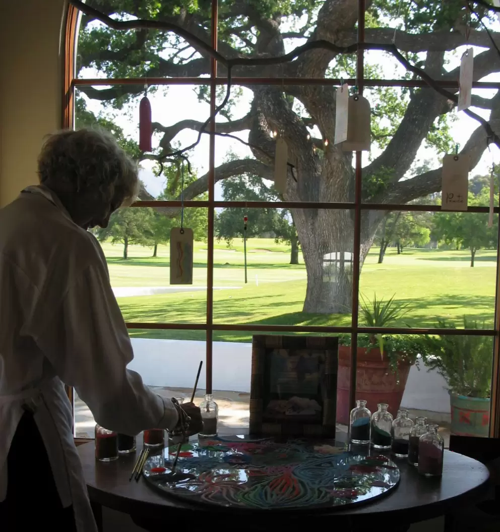 Renate, at the artist's cottage, painting patterns in colored sand with friendship tree behind. 