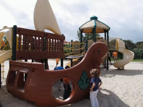 Wide-open beach with pale grey sand, shells on the shore, and aquamarine water, across from a playground, turtle center, and hike.