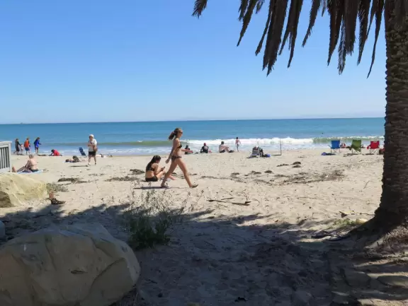 A crescent-shaped beach with rock pools, yellow and white cliffs, and surfers riding small waves near to shore. <br /><br />