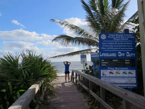 Wide-open beach with pale grey sand, shells on the shore, and aquamarine water, across from a playground, turtle center, and hike.