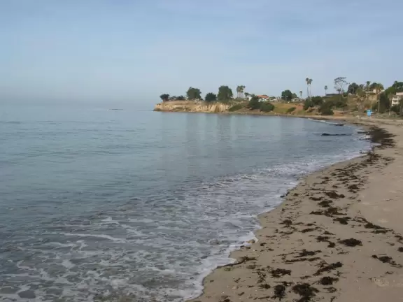 A crescent-shaped beach with rock pools, yellow and white cliffs, and surfers riding small waves near to shore. <br /><br />
