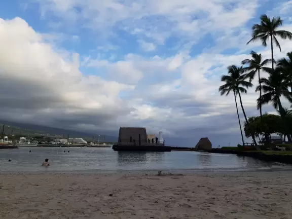 Pier with crystal clear water and views of the church steeple and high mountains backing the bay. Calm cove with Hawaiian hei'au out in the water.