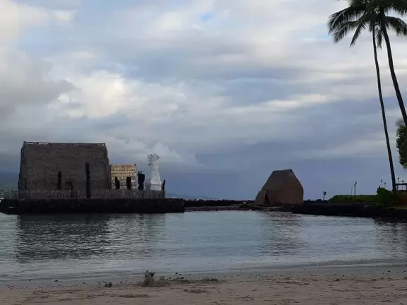 Pier with crystal clear water and views of the church steeple and high mountains backing the bay. Calm cove with Hawaiian hei'au out in the water.