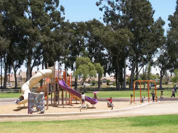 Large park with gazebo beside a lake with a fountain in the middle, pirate ship, manmade river, playground with huge slides, grassy hills galore, and pine trees.