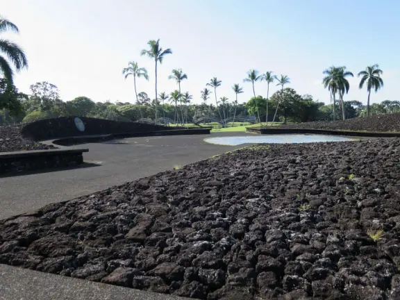 Beautiful statue backed by trees, with fruit and flower offerings below it. Huge lawn, narrow paths, picnic tables, and pedestrian bridge to Wailoa River State Park.