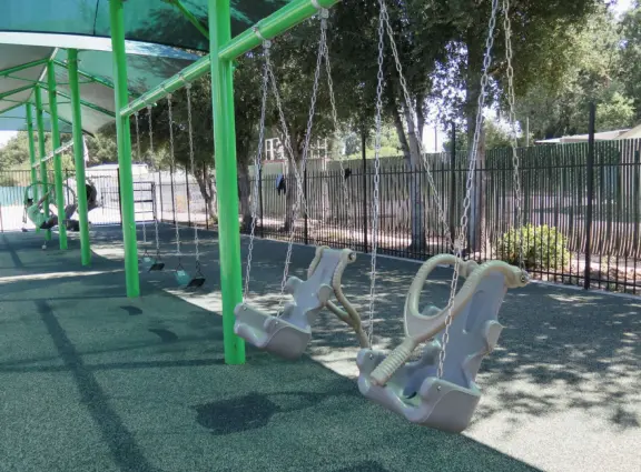Large, fenced, spread-out playground with treehouse structure, varied swings in the shade, dragonfly seesaw, egg seat, disc swing, and spinner.