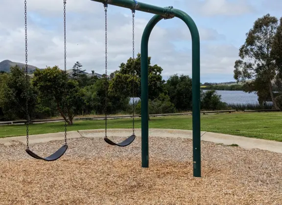Open space with trail through long dry grasses and views of the hills, beside a blue lake with geese, picnic tables, and a playground.