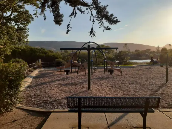 Open space with trail through long dry grasses and views of the hills, beside a blue lake with geese, picnic tables, and a playground.
